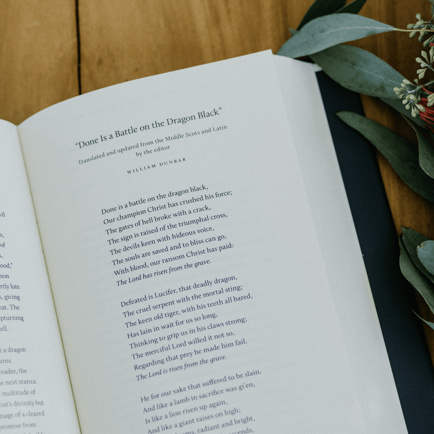 An open book showing the poem "Done Is a Battle on the Dragon Black" by William Dunbar on a wooden surface with green foliage nearby, promoting the book 100 Great Catholic Poems, book of Catholic poetry, Word on Fire, hardcover 9781685780746