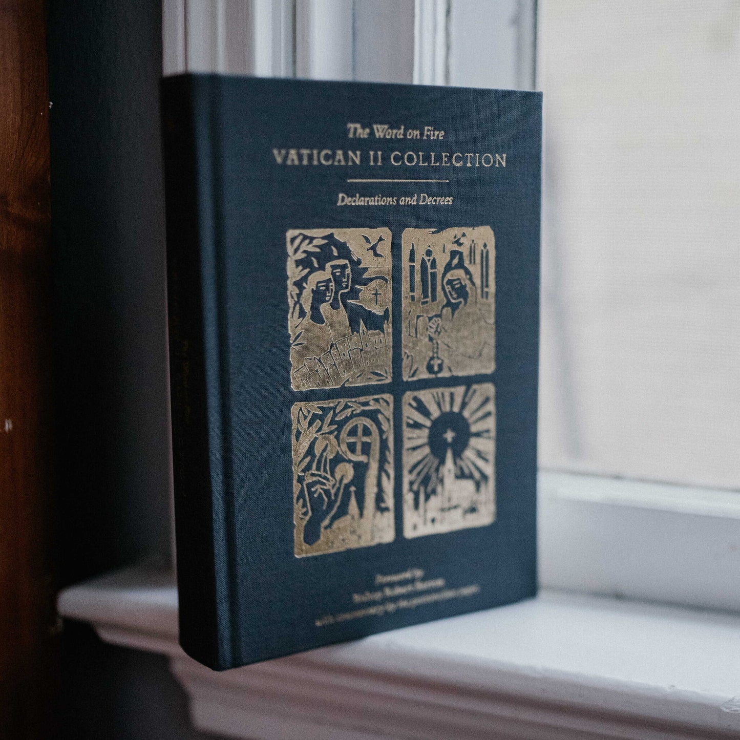 The Word on Fire Vatican II Collection: Declarations and Decrees book standing upright on a windowsill, featuring gold embossed religious imagery on a dark blue hardcover, with contributors indicated by Shopify metafields.